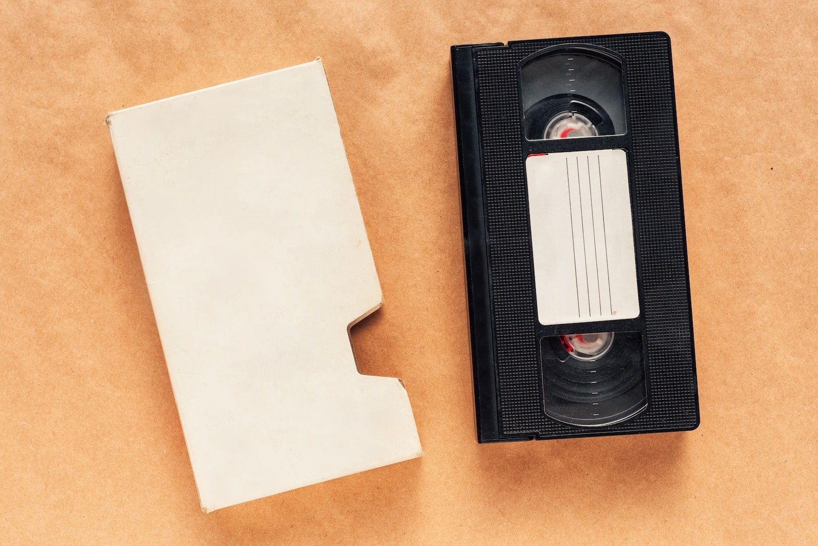 Image illustrant : Comment convertir des cassettes VHS en numérique ? A VHS tape with a blank label next to an empty white cardboard sleeve on a brown surface.