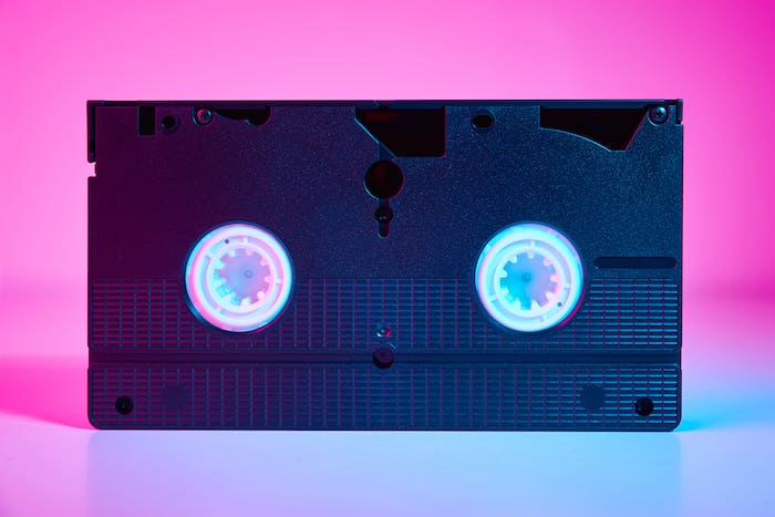 Image illustrant : Où faire numériser mes VHS ? A vintage VHS tape with visible reels, set against a vibrant pink and blue gradient background.