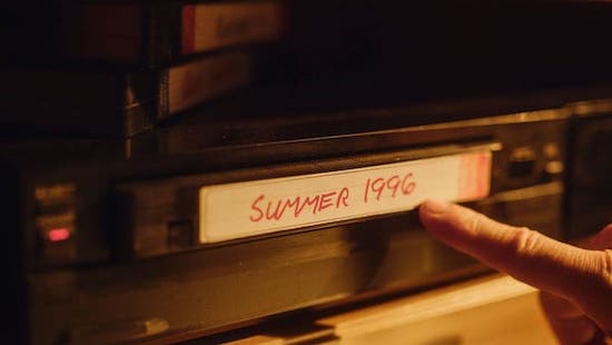 Image illustrant : Pourquoi numériser vos cassettes vidéo ? A hand points to a VHS tape labeled "Summer 1996" on a shelf, with other tapes stacked nearby.