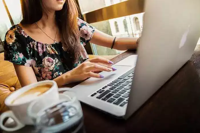 Image illustrant : Pourquoi numériser vos cassettes vidéo ? Woman typing on a laptop at a cafe table, with a floral top and purple nails. A cup of coffee and a glass of water are on the table.